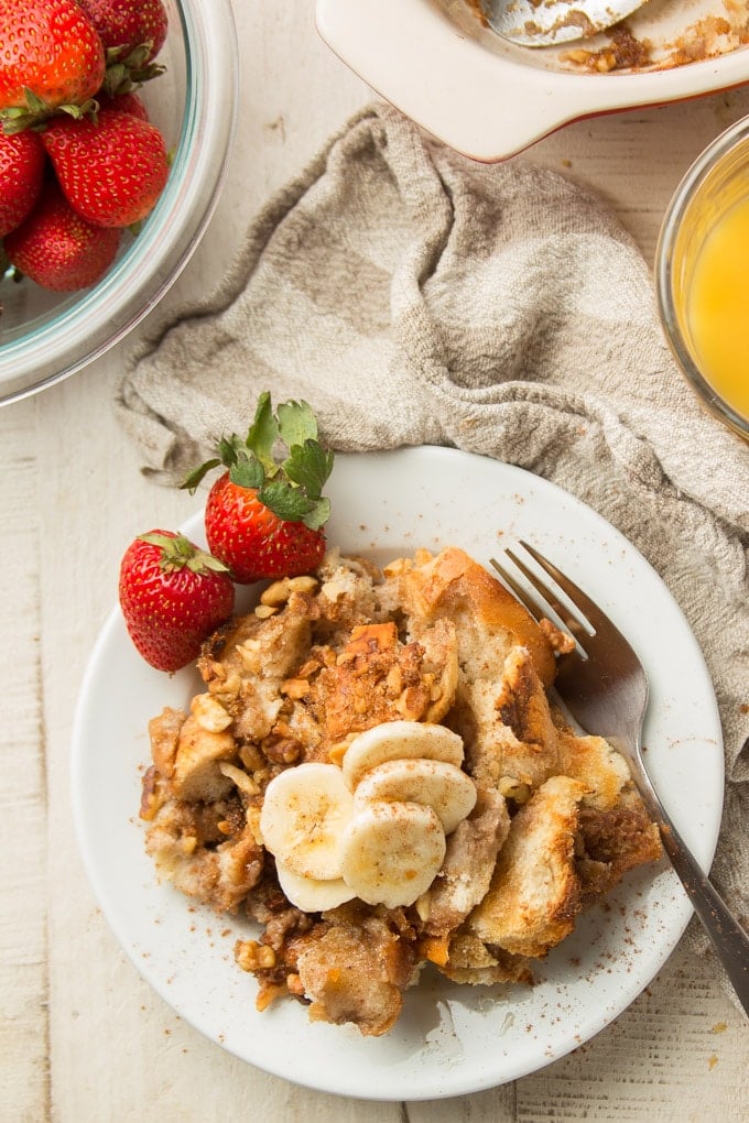 Overhead View of a White Wooden Table Set with Bowl of Strawberries, Glass of Orange Juice, and Piece of Vegan French Toast Casserole on a Plate