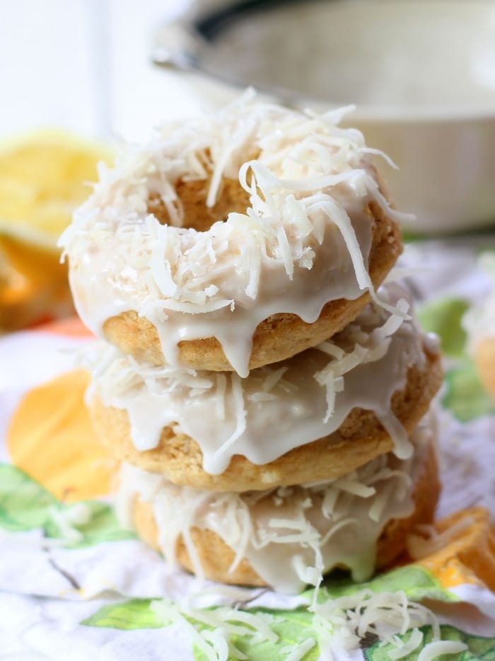 Stack of Three Lemon Coconut Doughnuts with Lemon Slices and Tea Cup in the Background