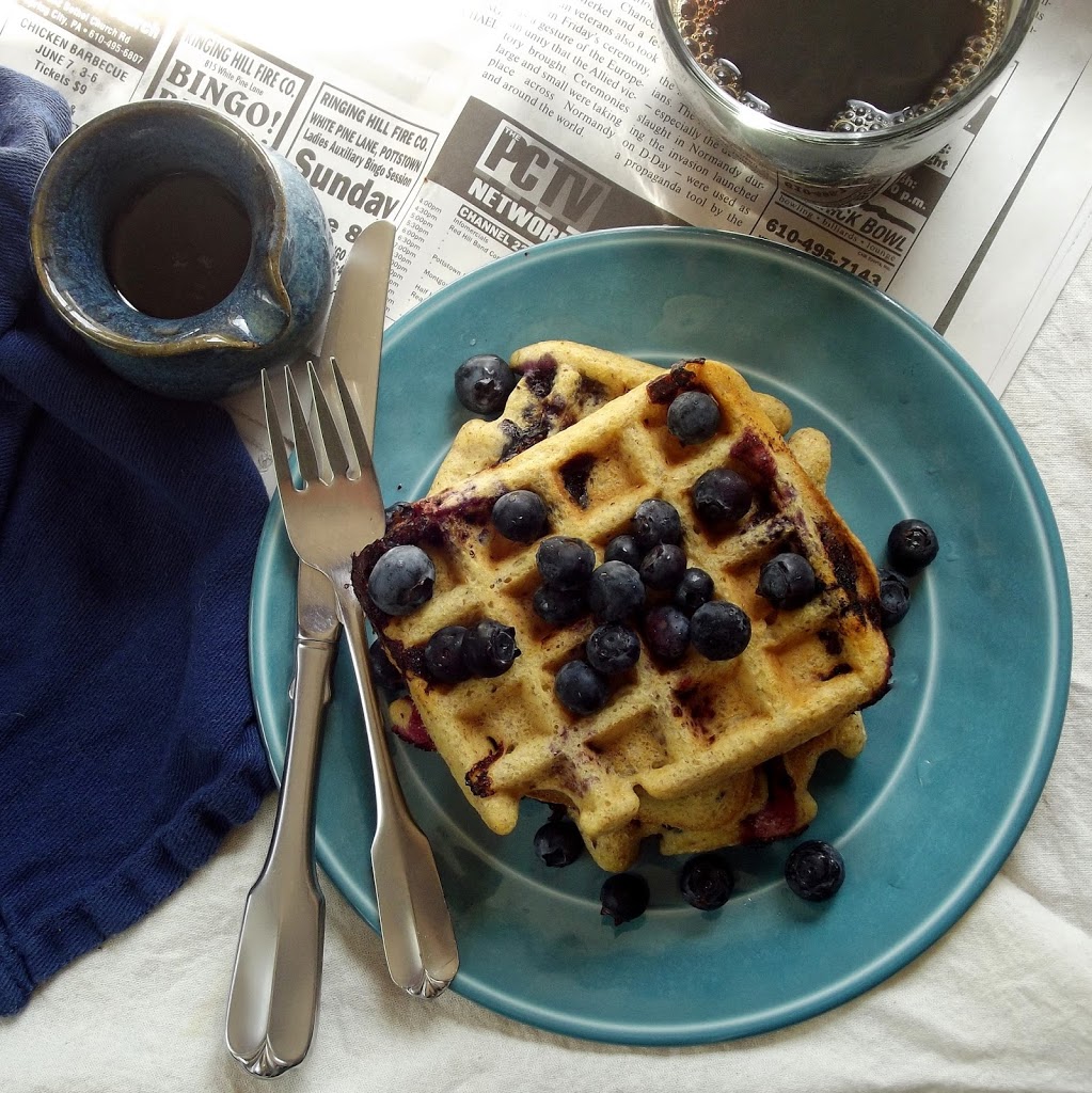 Overhead View of Blueberry Cornbread Waffles on a Plate with Fork and Knife, Jar of Maple Syrup and Coffee Cup on the Side