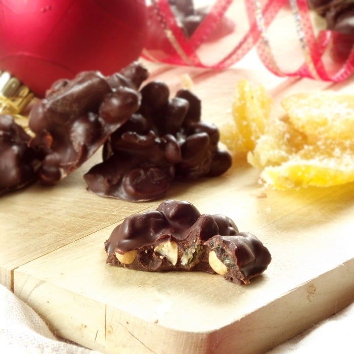 Close Up of a Peanut Ginger Cluster with a Bite Taken Out of It, with More Chocolates, Candied Ginger, and Christmas Ornaments in the Background