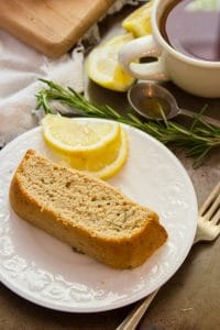 Slice of Rosemary Lemon Vegan Pound Cake on a Plate with Fork, Coffee Cup, Rosemary and Lemon Wedges in the Background