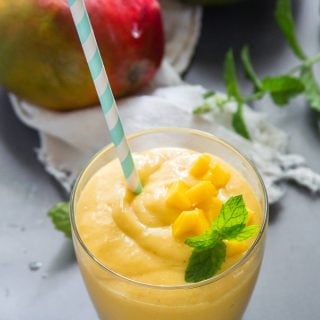 A Glass of Vegan Mango Lassi with Fresh Mint Leaves and Mangoes in the Background