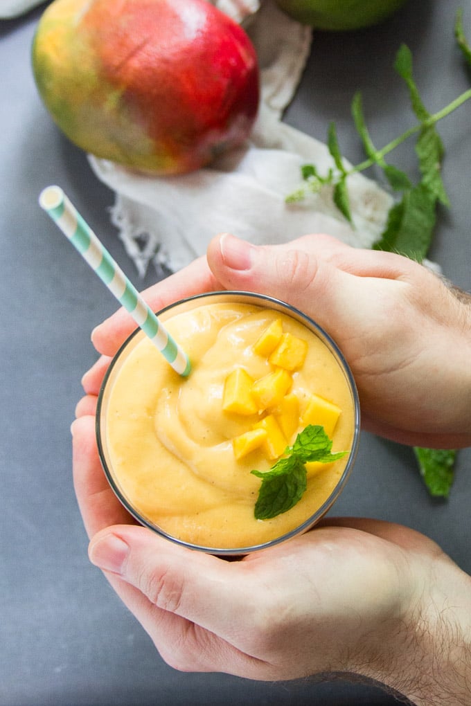 Pair of Hands Holding a Glass of Vegan Mango Lassi Over a Table