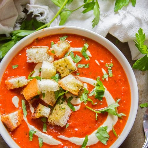 Overhead View of a Bowl of Overhead View of a Bowl of Roasted Red Pepper Soup with Garlic Bread Croutons with Spoon on the Side with Napkin and Parsley