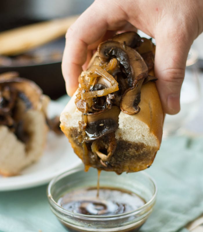 Hand Dipping Half of a Vegan French Dip Sandwich into a Bowl of Au Jus