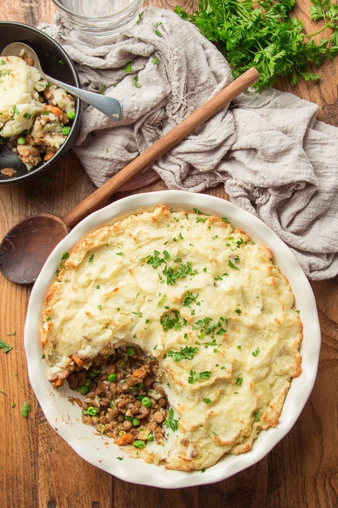 Wooden Surface Set with a Vegan Shepherd's Pie with Scoop Taken Out, Wooden Spoon, and Bowl Containing A Scoop of Vegan Shepherd's Pie