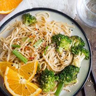 Plate of Spicy Orange Noodles and Broccoli with Orange Slices, Chopsticks, and Water Glass on the Side