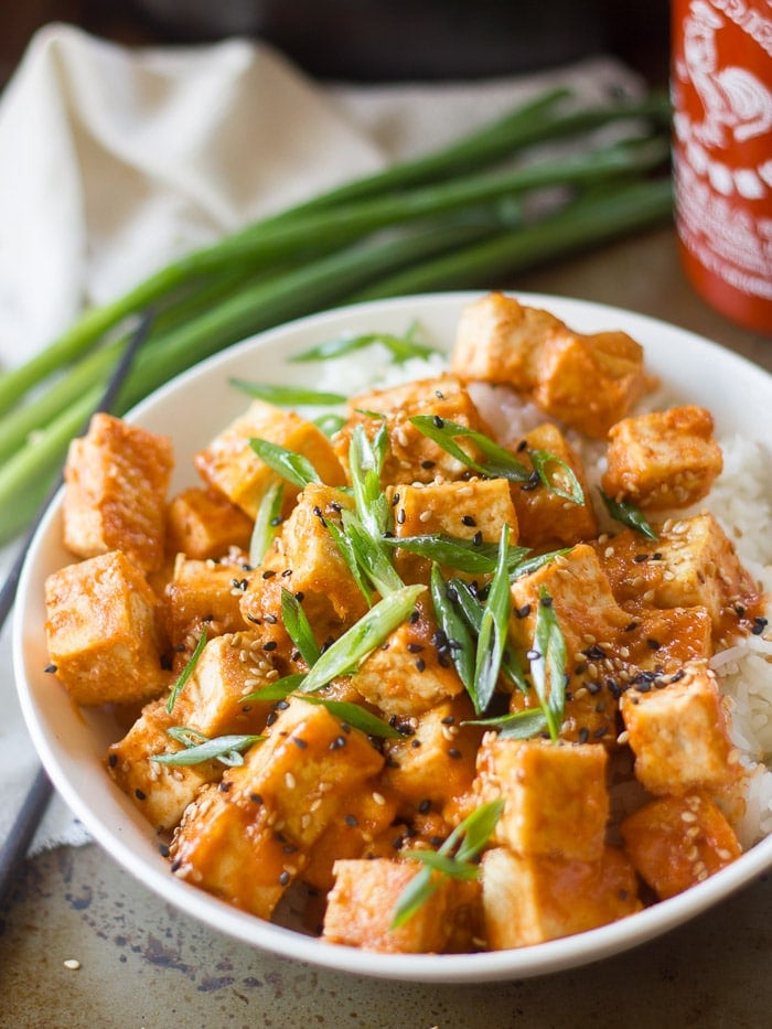 Bowl of Sesame Sriracha Tofu with Bunch of Scallions and Bottle of Sriracha in the Background
