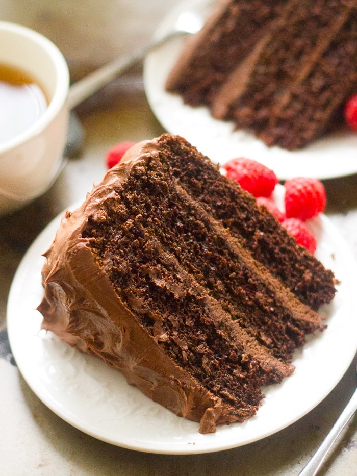 Slice of Vegan Mocha Layer Cake on a Plate with Raspberries, with a Second Slice of Cake and Tea Cup in the Background