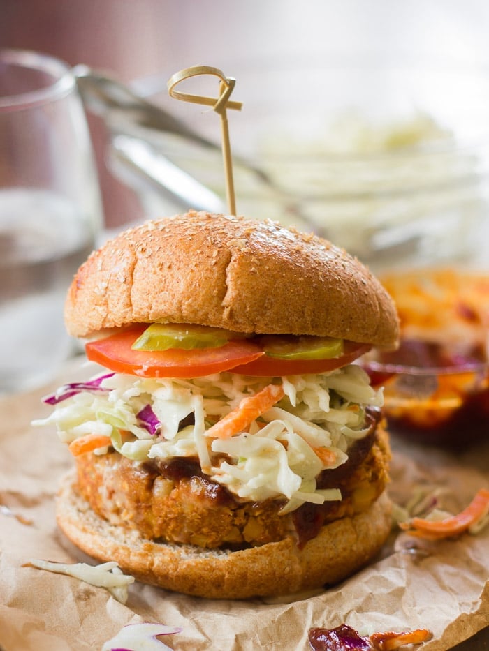 Barbecue Chickpea Burger on a Bun Topped with Slaw Barbecue Sauce and Tomato Slices with Glass Bowl and Drinking Glass in the Background.