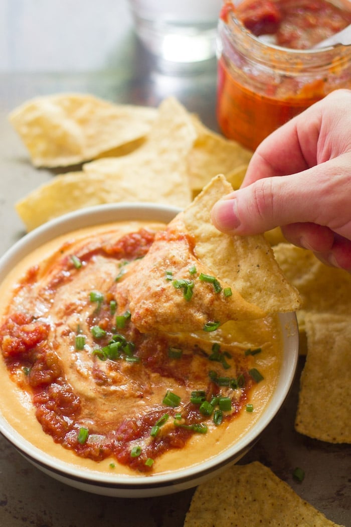 Hand Dipping a Chip into a Bowl of Nacho Cauliflower Cheese Topped with Salsa and Chives