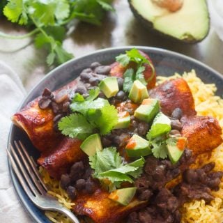 Three Black Bean Smothered Tofu & Plantain Enchiladas on a Plate Over Rice with Avocado, Cilantro, and Casserole Dish in the Background