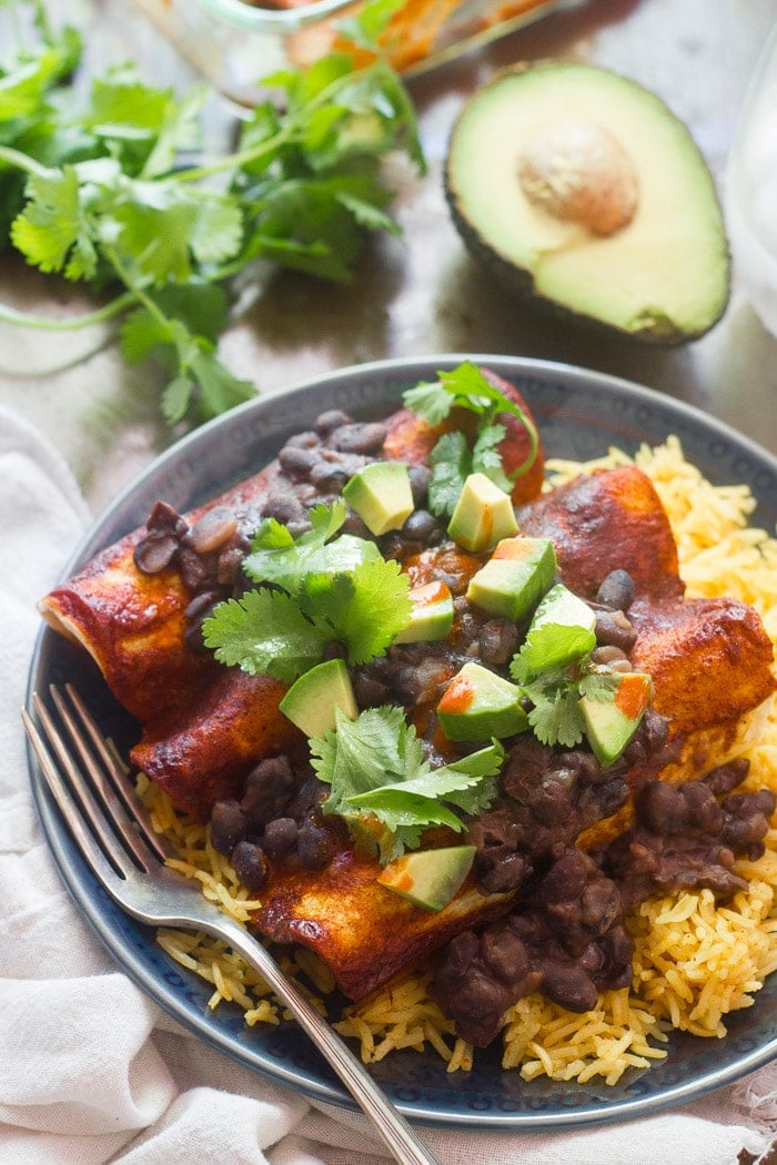 Three black bean smothered tofu and plantain enchiladas on a plate over rice with avocado, cilantro and casserole dish in the background.