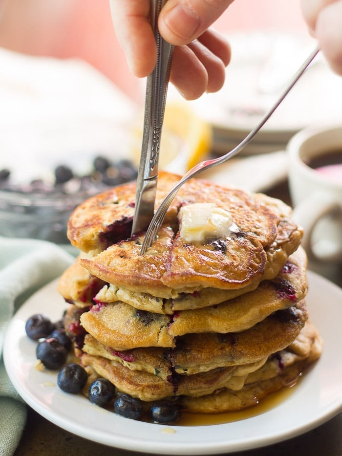Hand with Fork and Knife Cutting a Wedge into a Stack of Vegan Blueberry Pancakes