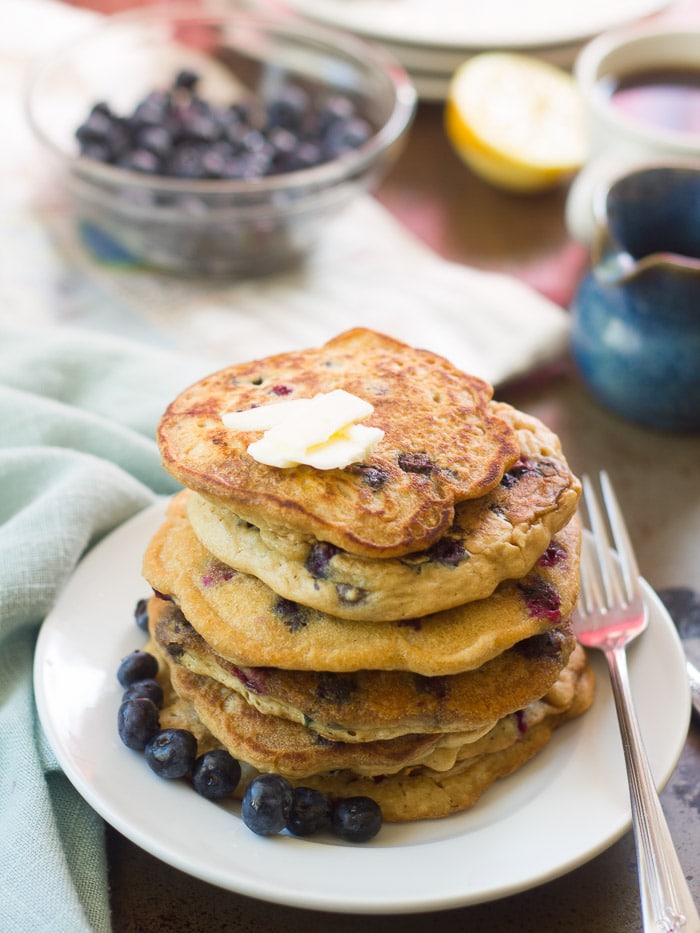 Stack of Vegan Blueberry Pancakes on a Plate with Bowl of Blueberries and Syrup Dispenser in the Background