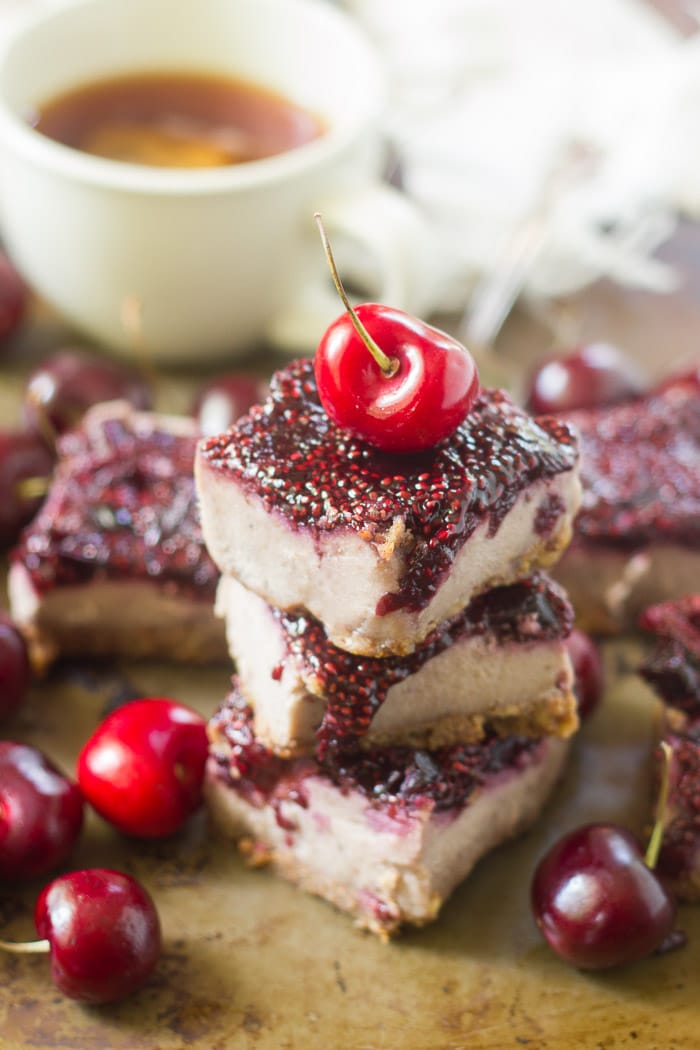 Stack of Three Vegan Cherry Cheesecake Bars with More Bars, Cherries, and a Teacup in the Background
