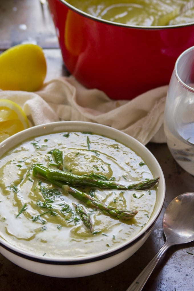 A Bowl of Vegan Cream of Asparagus Soup with a Red Pot and Lemon Wedges in the Background