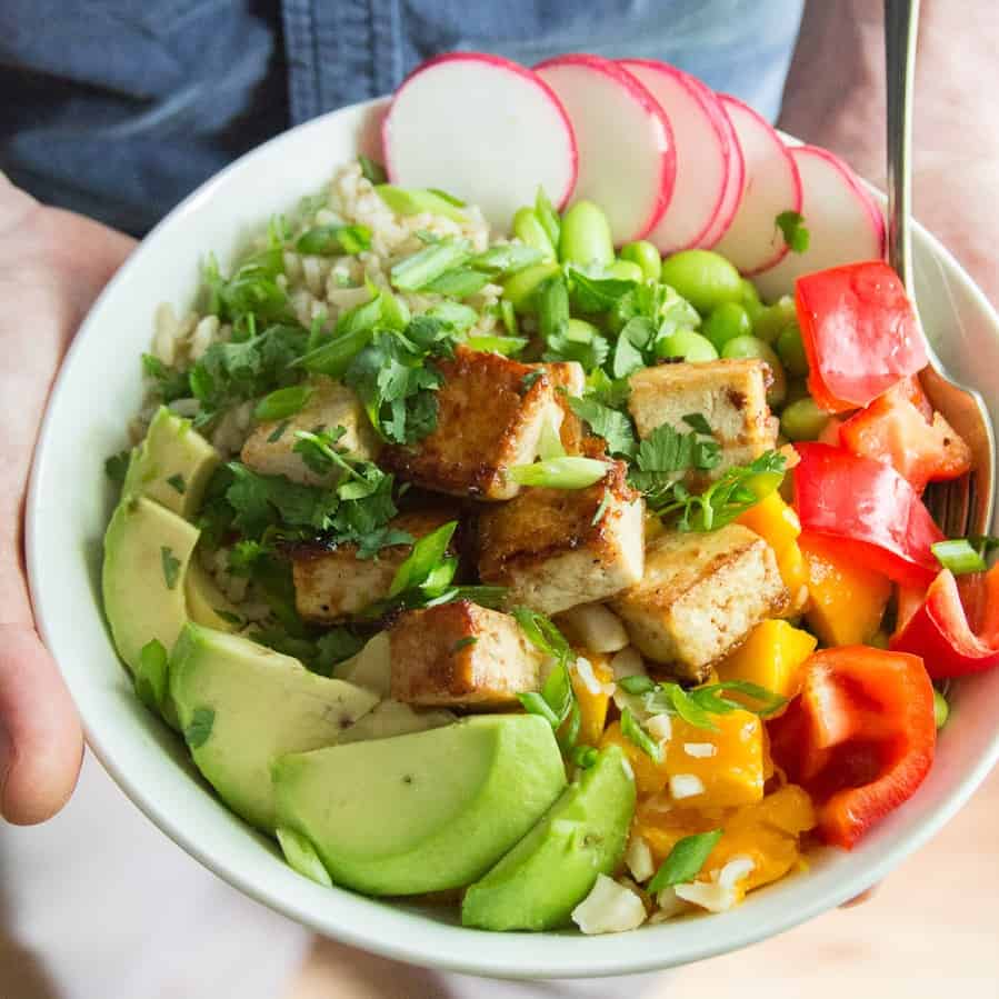 Hands Holding a vegan poke bowl with tofu, vegetables and avocado slices.