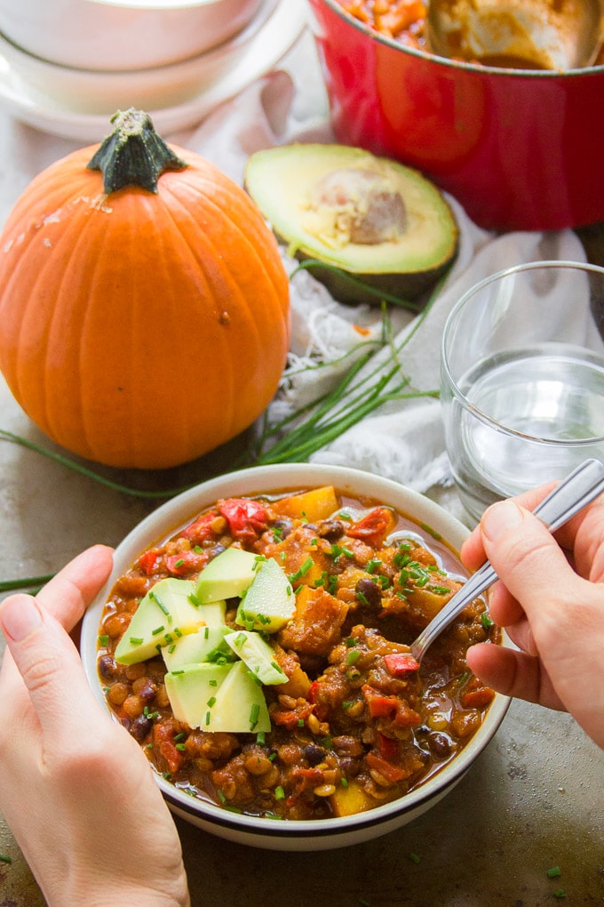 Hands Holding a Bowl of Triple Pumpkin Chili with Spoon Over Table Set with Water Glass and Pumpkin