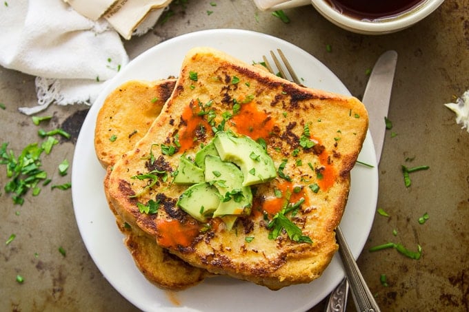 Overhead View of Two Slices of Savory Vegan French Toast Topped with Avocado Slices, Herbs and Hot Sauce on a Plate