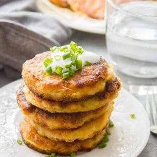 Stack of Cheesy Vegan Mashed Potato Pancakes on a Plate with Fork, Drinking Glass and Napkin in the Background