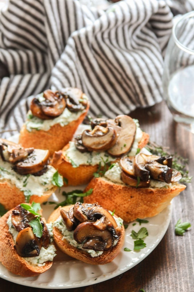 Mushroom Crostini with Herbed Cashew Cheese on a Plate with Drinking Glass in the Background