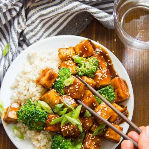 Chopsticks Grabbing a Piece of Tofu From a Plate of Teriyaki Tofu with Broccoli