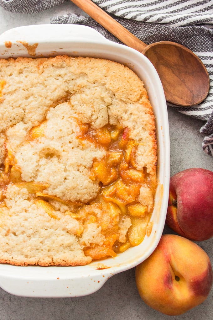 Vegan Peach Cobbler in a Baking Dish Surrounded by Fresh Peaches and a Wooden Spoon