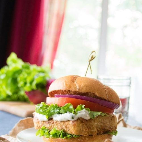Crispy Tofu "Fish" Sandwich with Lettuce, Tomato and Onion on a Plate with Water Glass in the Background