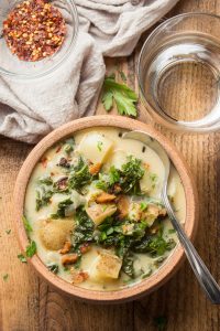 Vegan Zuppa Toscana in a Clay Bowl with Spoon, Water Glass and Bowl of Red Pepper Flakes