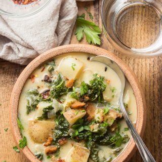 Vegan Zuppa Toscana in a Clay Bowl with Spoon, Water Glass and Bowl of Red Pepper Flakes