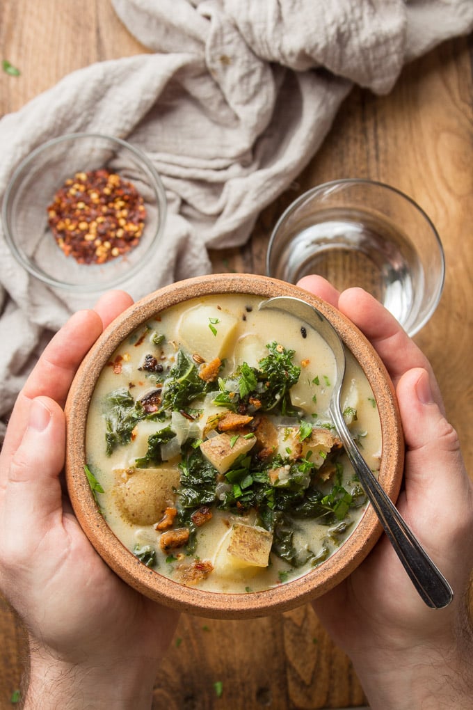 Pair of Hands Holding a Bowl of Vegan Zuppa Toscana Over a Wooden Table