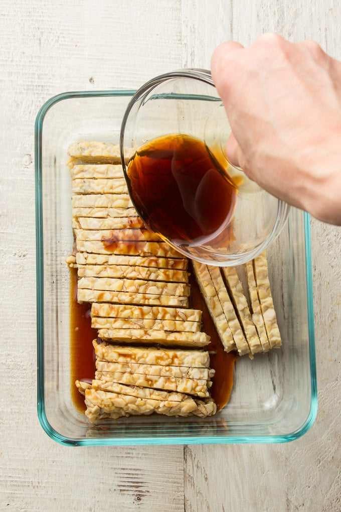 Hand Pouring Marinade Over Tempeh in a Glass Dish