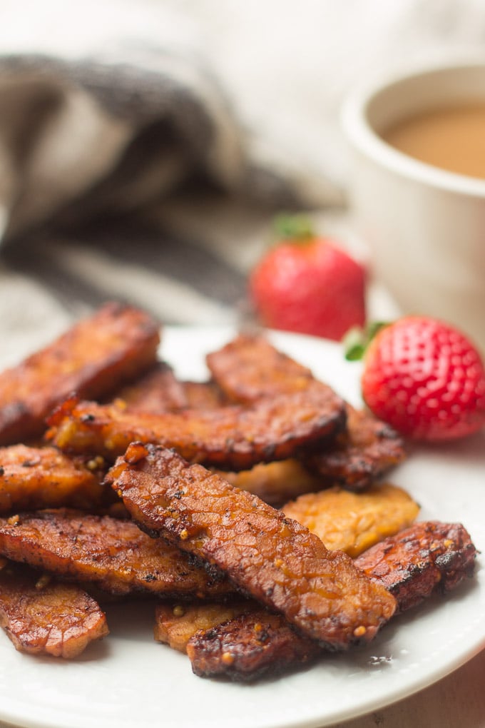 Close Up of Tempeh Bacon Slices on a Plate with Strawberries and Coffee Cup in the Background