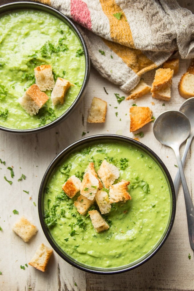 Two Bowls of Fresh Pea Soup on a White Wooden Table with Spoons