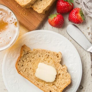 White Wooden Surface Set with Slices of Beer Bread on a Cutting Board, Slice of Beer Bread on a Plate, Glass of Beer, and Three Strawberries