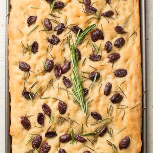 Loaf of Olive Rosemary Focaccia Topped with a Sprig of Fresh Rosemary on a White Wooden Surface