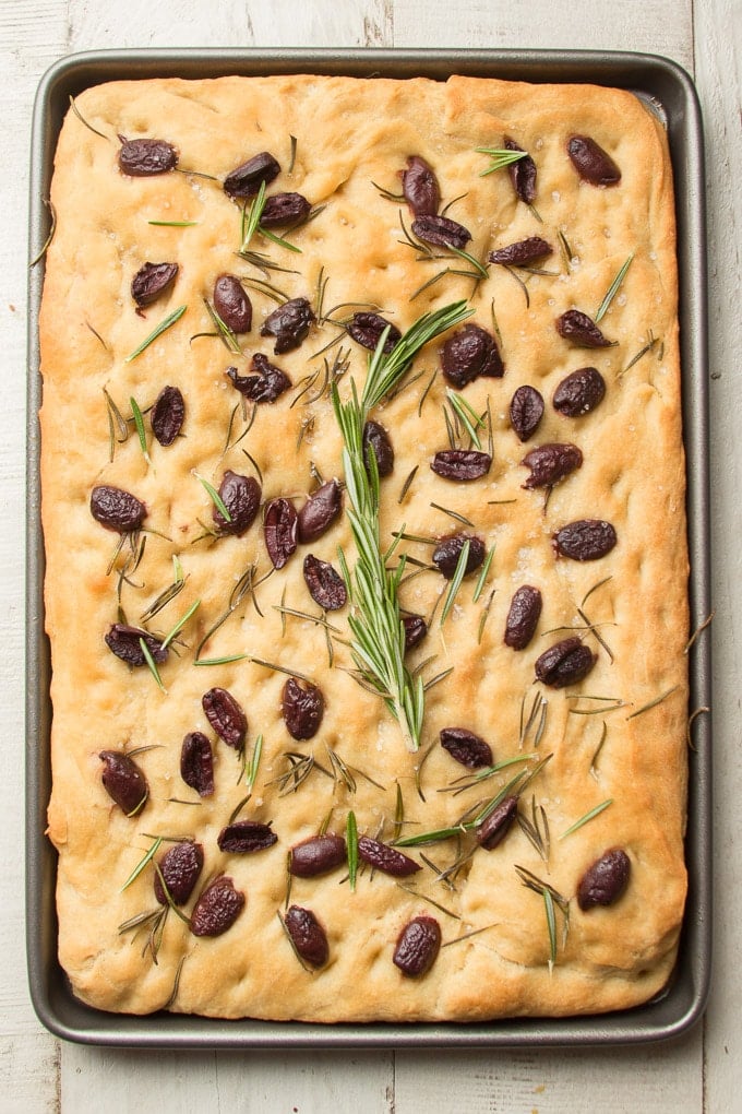 Loaf of Olive Rosemary Focaccia Topped with a Sprig of Fresh Rosemary on a White Wooden Surface