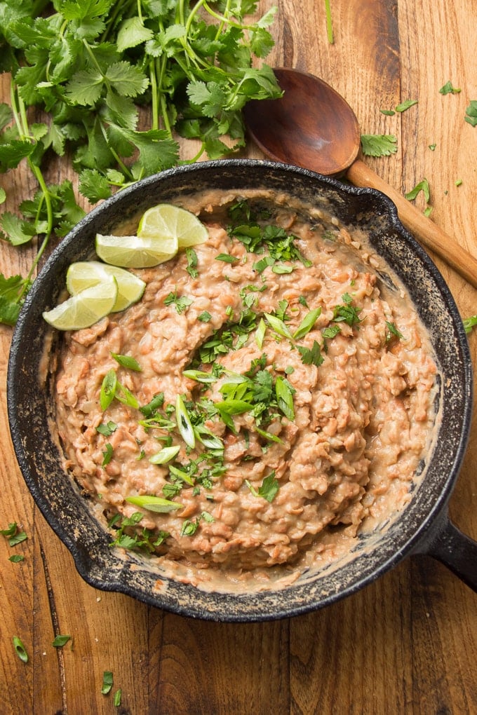 Skillet of refried beans on wooden surface with spoon and bunch of cilantro.