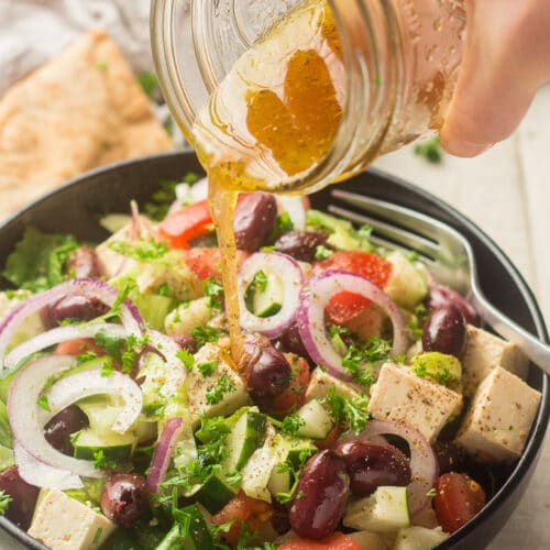 Hand Pouring Dressing Over Vegan Greek Salad in a Serving Bowl