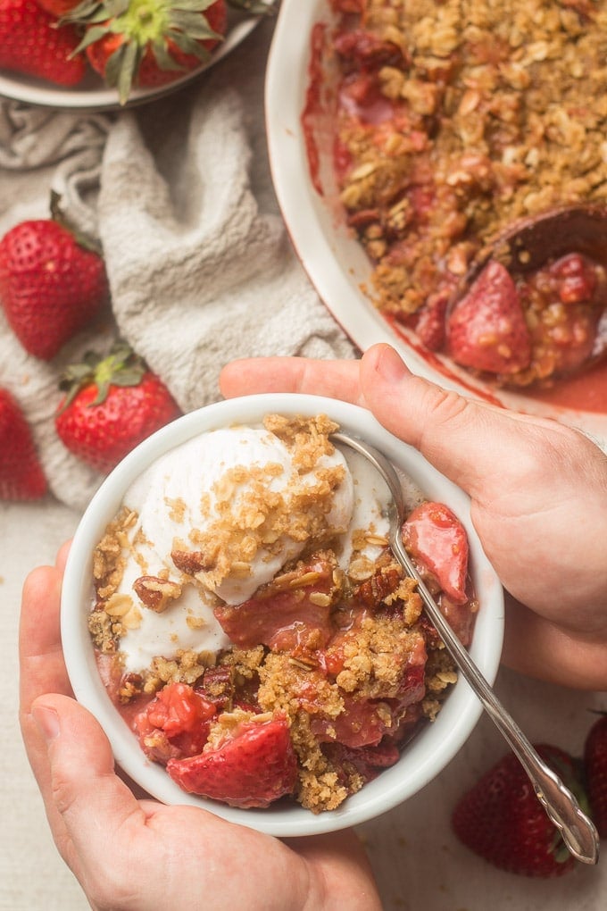 Pair of Hands Holding a Bowl of Vegan Strawberry Crisp and Vanilla Ice Cream Over a Table