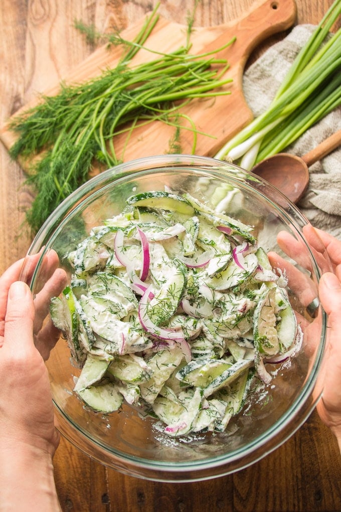 Pair of Hands Holding a Bowl of Creamy Cucumber Dill Salad Over a Table