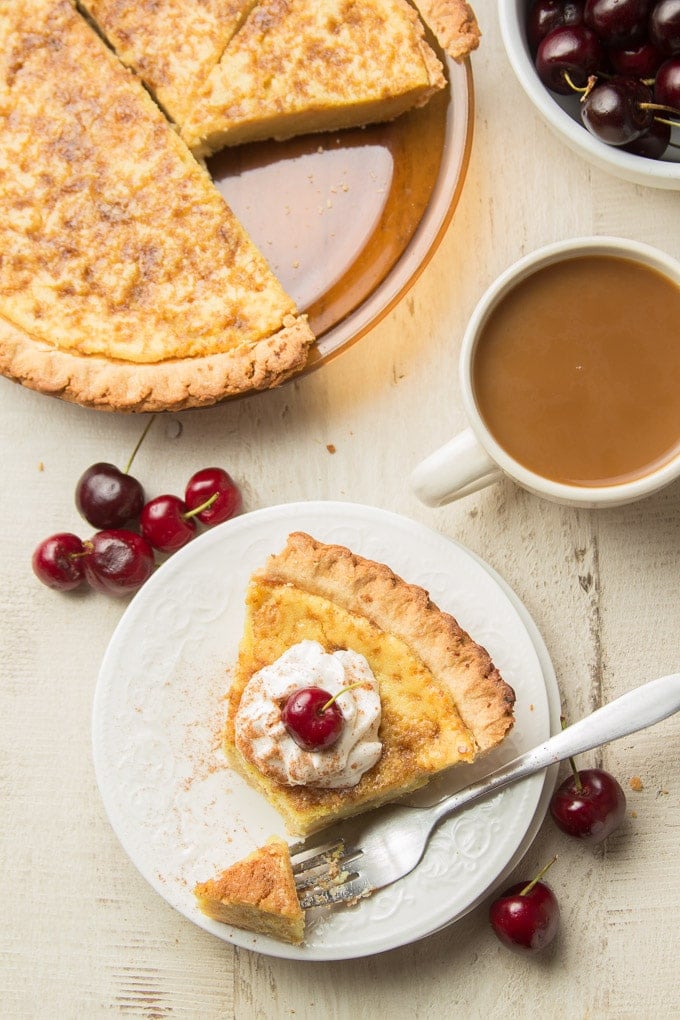 Overhead View of a White Table Set with Pie Plate, Bowl of Cherries, Coffee Cup, and Slice of Vegan Custard Pie with Whipped Topping an Cherry