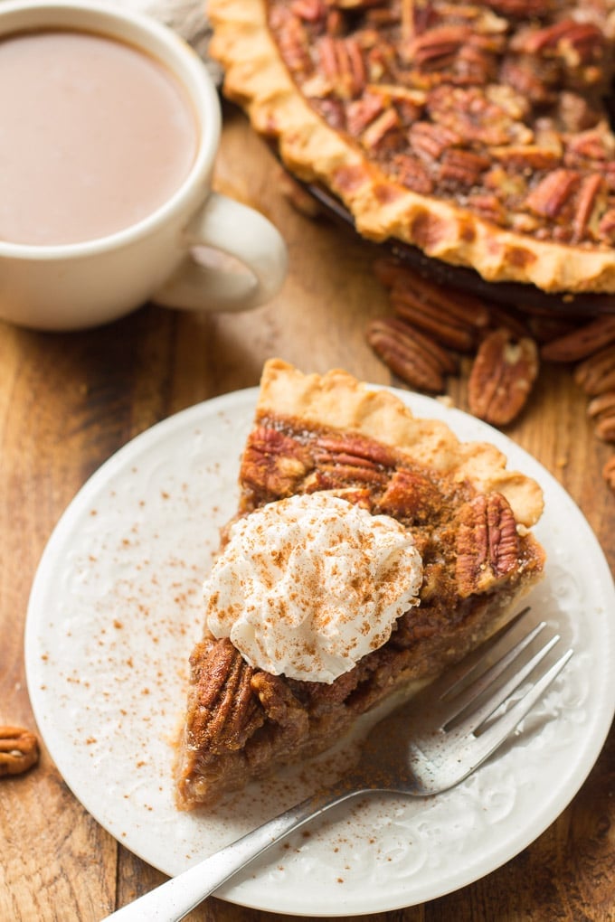 Slice of Vegan Pecan Pie with Whipped Topping on a Plate, with Coffee Cup and Rest of Pie in Background