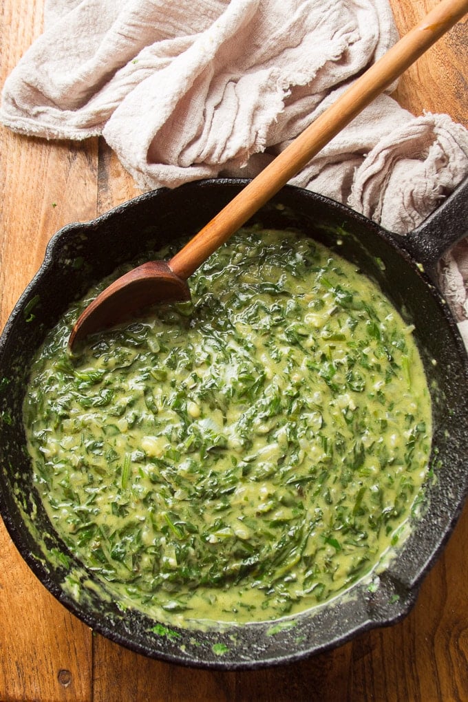 Cast Iron Skillet Sitting on a Wood Surface and Filled with Vegan Creamed Spinach with Wooden Spoon