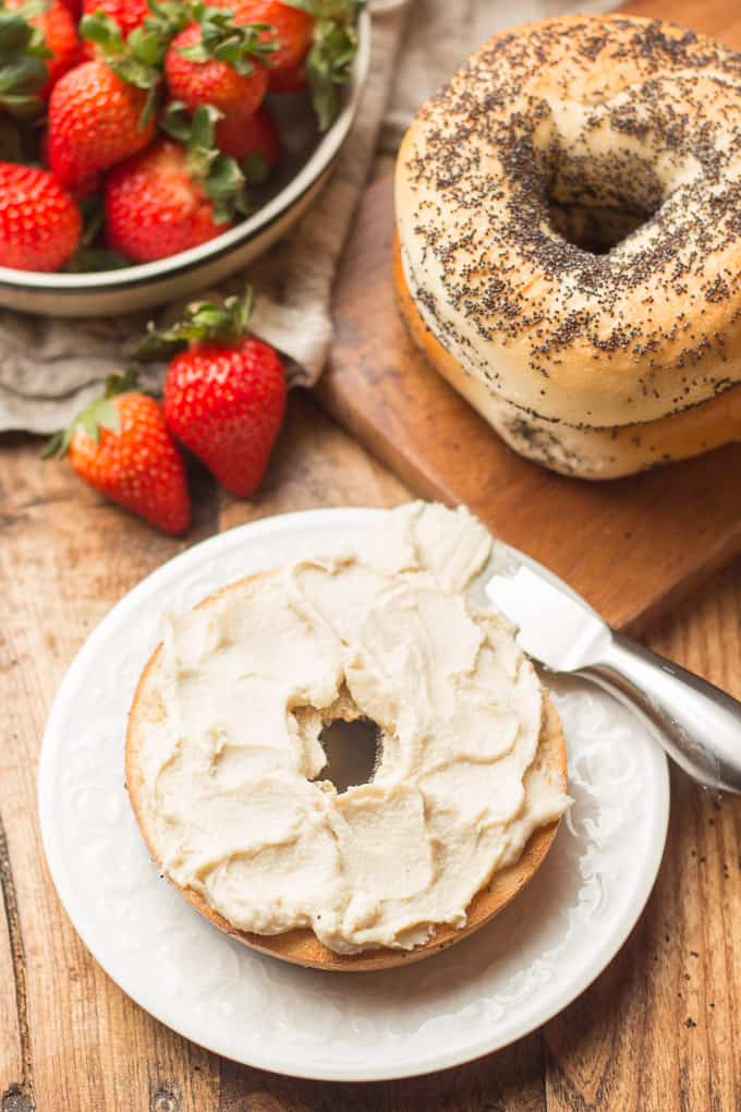 Half of a Bagel on a Plate with Vegan Cream Cheese on Top, Strawberries and Whole Bagels in the Background