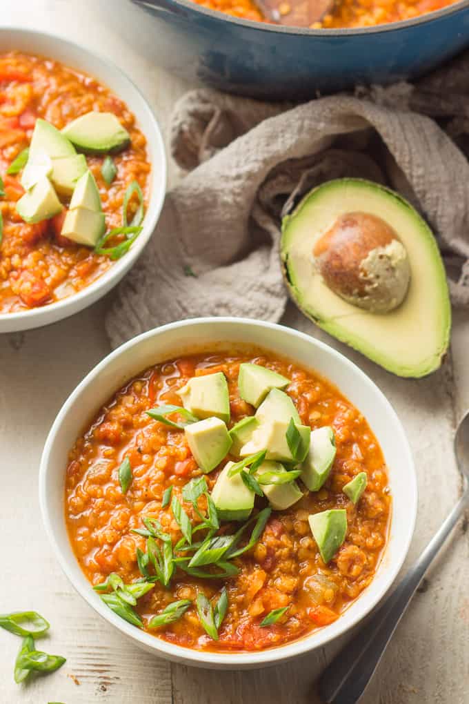 Two Bowls of Red Lentil Chili, Avocado Half, Spoon and Tea Towel on a White Wooden Surface