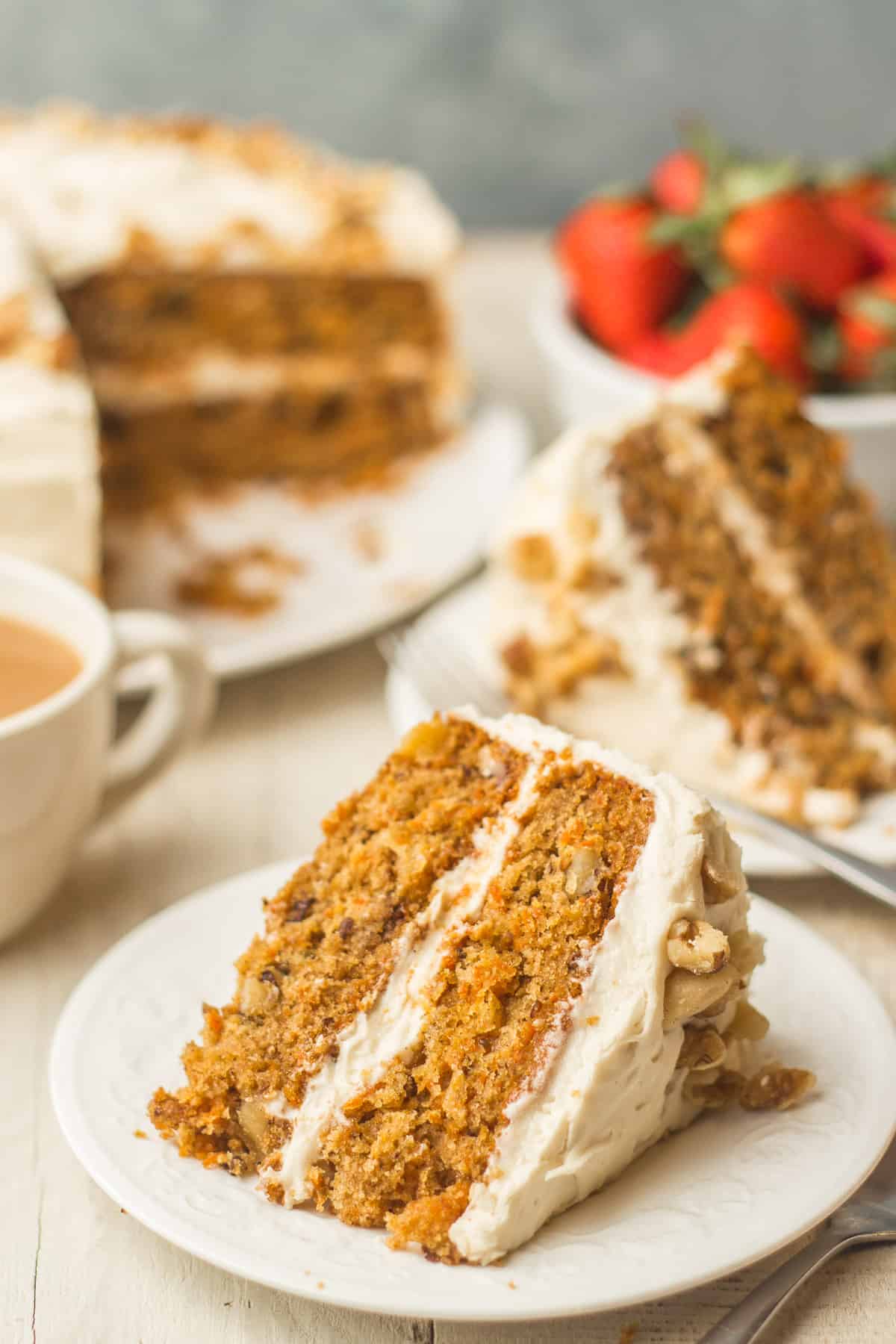 Slice of Vegan Carrot Cake on a Plate with Additional Slices, Coffee Cup and Strawberries in the Background