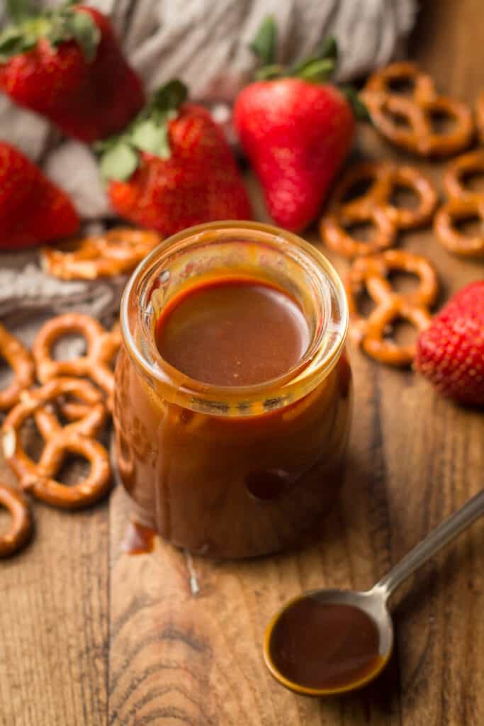 Jar of Vegan Caramel Sauce Surrounded By Strawberries, Pretzels and Spoon