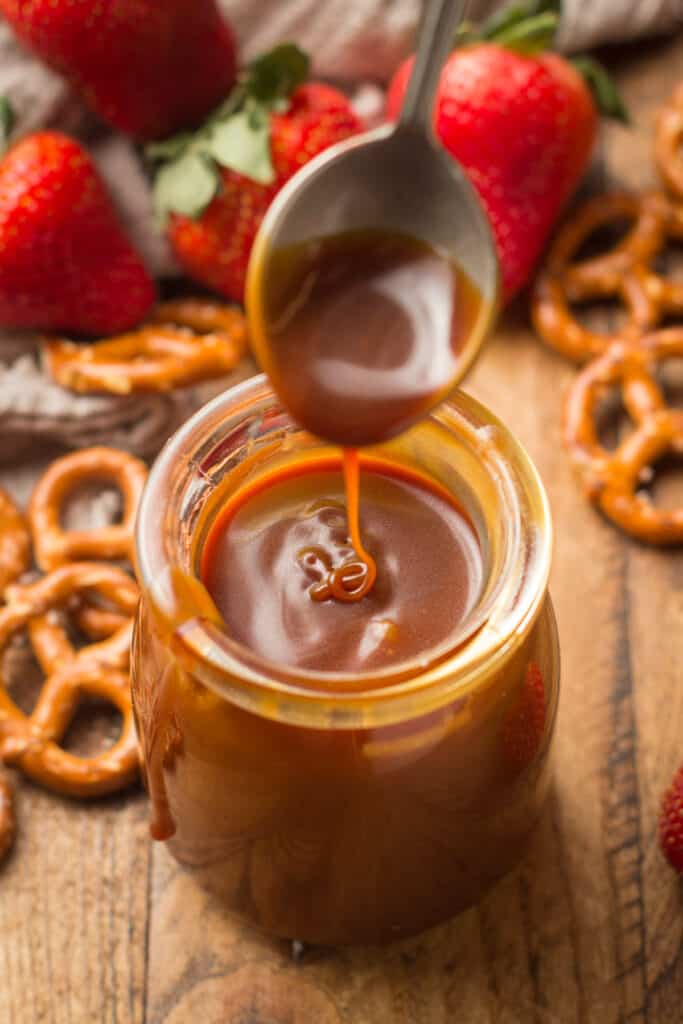 Close Up of Spoon Drizzling Vegan Caramel Sauce into a Jar with Strawberries and Pretzels in the Background